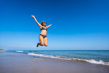 Joyful summer. Young woman wearing black and white bikini and sunglasses jumping on sandy beach against blue sky. Energy, happiness and activity.