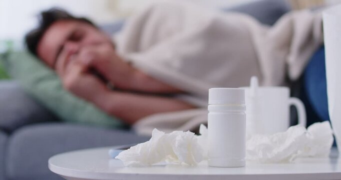 Sick young man lying on sofa at home with seasonal flu or cold feeling unhealthy with influenza and runny nose. Male person suffering from cough with a pills and napkins on table. 4k video.