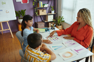 Teacher sitting with two students at table, engaging in alphabet letter activity. Bookshelf and large window visible in classroom setting, plants adding to a cozy environment
