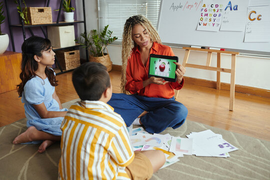 Teacher engaging group of young children in learning alphabet displayed on tablet surrounded by colorful educational tools in lively classroom environment
