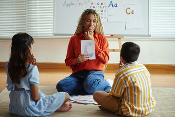 Smiling teacher showing letter C to two children during alphabet lesson. Sitting on classroom floor, children attentive and participating actively