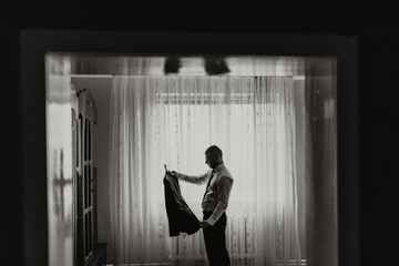 A man is standing in front of a mirror, putting on a jacket. The image has a moody and somewhat...