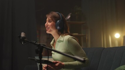 Footage of beautiful online radio host using professional microphone while recording podcast in living room. Confident young Caucasian woman speaking to radio listeners. Evening time. Appartement