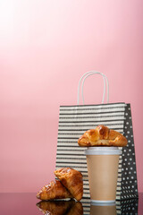 vertical view of disposable coffee cup, croissant and shoping bag with pink background