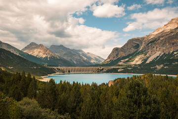 The tranquil waters of Lake Cancano, Valdidentro, Italy surrounded by lush greenery and majestic mountains, with the St. James Dam visible in the background under a partly cloudy sky.