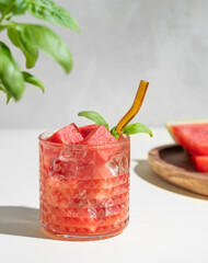 Glass of refreshing watermelon cocktail with basil and ice with fruit slices on  a light background with shadow and fresh herb.