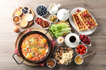 Tasty shakshouka in frying pan and other meals served for breakfast on wooden table, flat lay
