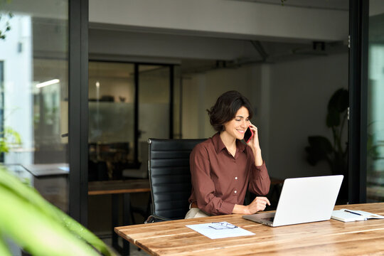 Busy happy young woman manager talking on phone in office. Professional lady entrepreneur, business woman client manager making business call on cellphone at work communicating using laptop in office.
