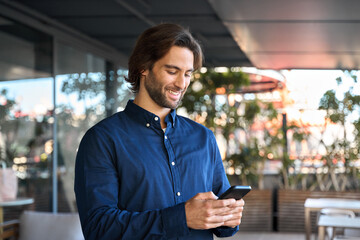 Happy young business man using mobile phone standing outdoors. Smiling guy holding smartphone using...