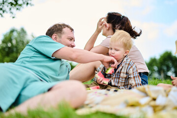 Happy Caucasian family on picnic. Mom, dad, son and daughter sitting on picnic blanket on green grass in yard, eating watermelon and having fun