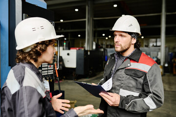 Industrial workers in protective gear discussing operational procedures while checking notes in an industrial workshop environment