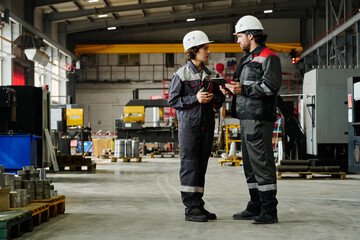 Two industrial workers in safety gear discussing work tasks inside large warehouse with machinery and equipment
