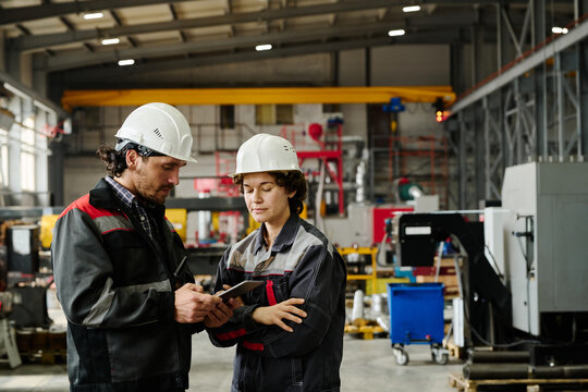 Engineers wearing safety helmets collaborating on project in industrial workshop with various machinery in background