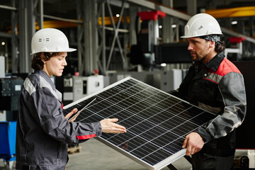 Two workers wearing hard hats and work uniforms standing in factory setting, carefully examining solar panel