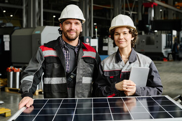 Engineers wearing hard hats and safety jackets standing next to a solar panel, smiling in industrial factory