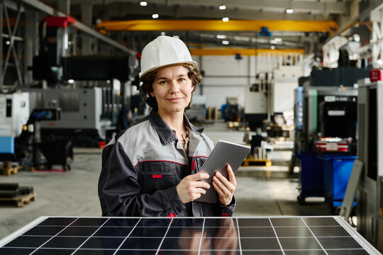 Portrait of female engineer standing in large industrial workshop holding tablet while smiling at camera