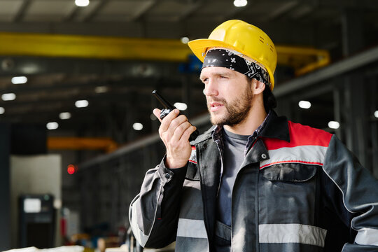 Mechanic holding walkie talkie while standing indoors in industrial setting and wearing a yellow hard hat and protective gear