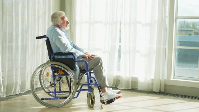 Sad senior man sitting in wheelchair looking out window in retirement home hospital. Old paralyzed man in chair for people with disability feel depressed. Lonely elderly sick man sitting on wheelchair