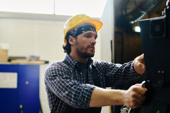 Man wearing a yellow hard hat and checkered shirt adjusting industrial equipment in a factory
