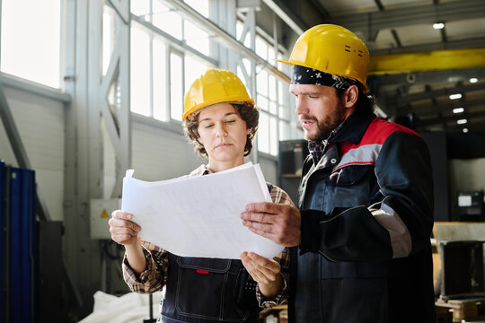 Two engineers reading construction plans in an industrial workshop while wearing safety helmets