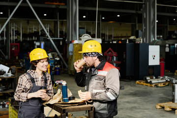 Employees standing in workshop area, taking break with coffee cups, wearing hard hats and work uniforms