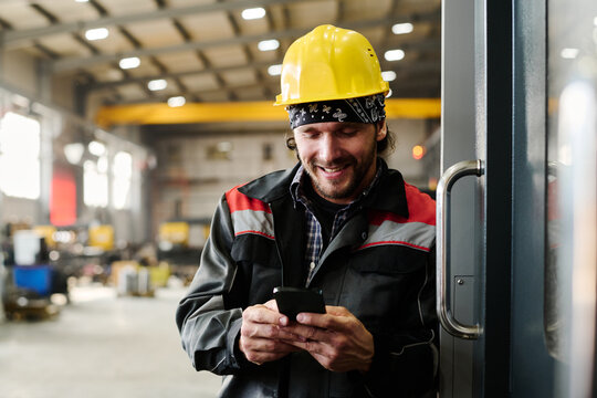 Smiling factory worker wearing yellow hard hat and black jacket checking phone