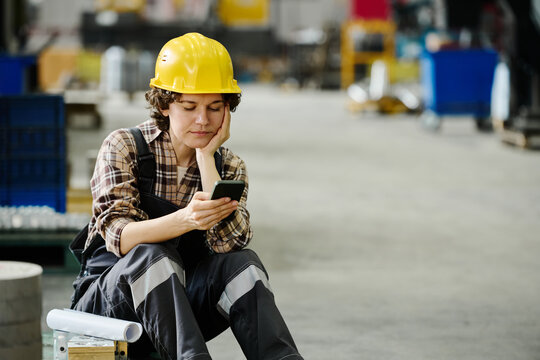 Industrial worker wearing protective gear sitting and checking smartphone in warehouse setting, appearing thoughtful and engaged in activity