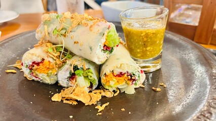 Close-up, salad, roll, and salad dressing placed on a black plate, a wooden table in a restaurant. Vegetable chicken rolls salad with Thai sauce. Fresh Vietnamese spring rolls on a plate with dipping.