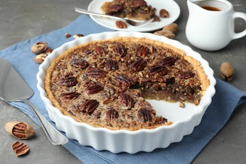 Delicious pecan pie in baking dish, cake server and fresh nuts on gray textured table, closeup