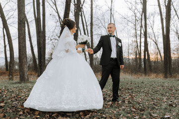 A bride and groom are walking through a forest, hand in hand. The bride is wearing a white dress and the groom is wearing a black suit. Scene is romantic and peaceful