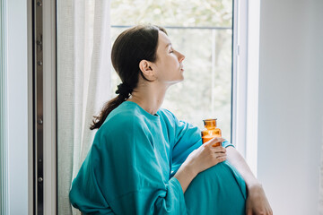 Woman in blue sitting by the window holding candle, practicing autumn fall self-care, mindfulness,...