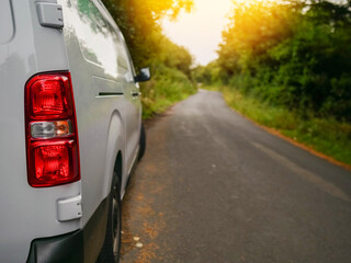 A white medium size van is parked on a small county road in rural area with a red tail light. The...