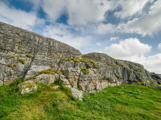 A rocky hillside with a grassy area in between. The sky is cloudy and the grass is green. Burren, Ireland. Famous Irish landscape and popular busy tourist area. Warm sunny day.