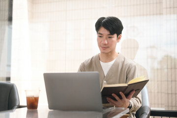 A Young adult man combines traditional reading with modern technology, using a laptop while holding a book, symbolizing the blend of digital and analog learning methods