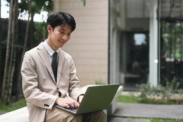 A young businessman in a beige suit and tie sits outdoors, smiling as he works on his laptop, demonstrating a blend of productivity and enjoyment in an outdoor serene