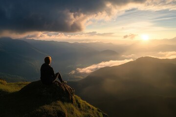 Obraz premium A person sits on a rock, observing the serene sunrise over a vast mountain range, with clouds beneath and a golden light breaking through, capturing a moment of tranquility.