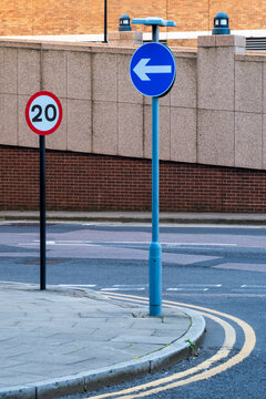 This image features a close-up of traffic signs in an urban setting, with a 20 mph speed limit sign and a blue directional arrow sign, along with road markings and a sidewalk curb.