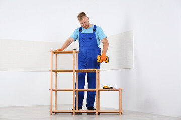 Worker with electric screwdriver assembling wooden shelving unit indoors