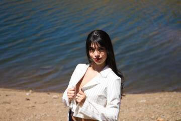 Portrait of a beautiful young brunette latin woman with red lips, wearing a white shirt and black shorts, holding her shirt with her hands. In the background a lake with crystal clear blue water.