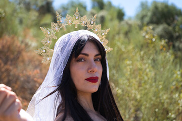 beautiful young brunette latin woman with red lips and dressed with a white veil and a golden crown of virgin mary or goddess. The woman is in a forest with vegetation as if she were an apparition.