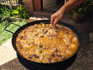 A close-up of a person stirring a traditional Spanish paella in a large pan outdoors.