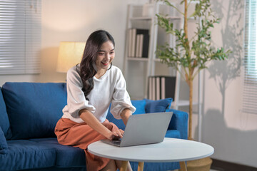 Young asian woman smiling and typing on laptop while working from home on sofa