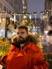 Fototapeta premium Handsome millennial man model with a hairstyle in winter red jacket walks in night city with festive lights. Christmas and winter holidays season