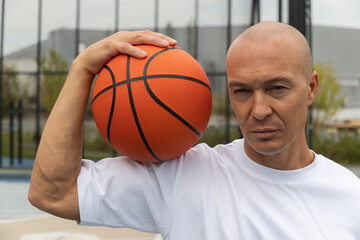 Portrait of sporty man holding basketball orange ball on shoulder and looking at camera on sports court. Close up. Outdoors.