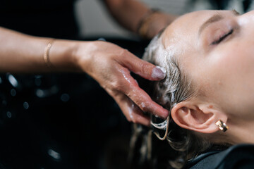 Closeup hands of hairdresser washing head of young woman in beauty hairdressing salon. Close-up of unrecognizable hairstylist foaming shampoo on long blonde hair of beautiful female client.