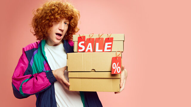 Sales in full swing. Surprised young man with curly hair holding stack of boxes after online shopping against pink studio background. Concept of Black Friday, Cyber Monday, fashion, Flash Sale. Ad - Powered by Adobe