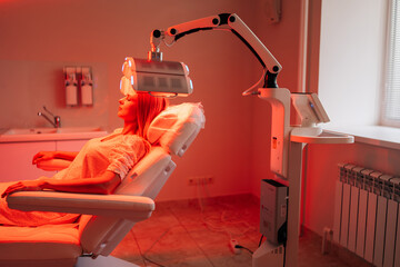 Attractive young woman having non-invasive type of phototherapy in medical clinic during hair treatment. Blonde female lying on chair closed eyes having LED light hair photodynamic therapy.
