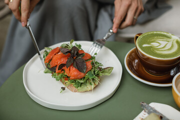A woman enjoying a delicious breakfast of toast with salmon, avocado, and herbs with a cup of matcha latte on the terrace.