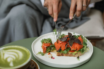 A woman enjoying a delicious breakfast of toast with salmon, avocado, and herbs with a cup of matcha latte on the terrace.