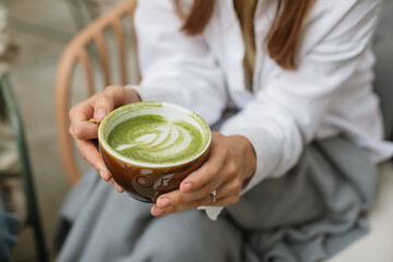 A close-up of a young woman holding a cup of matcha latte sitting on a cafe terrace.